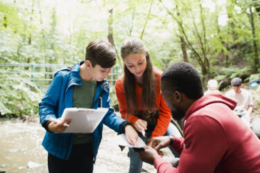 Two school children documenting and comparing their findings on the field trip with each other. They are getting advice from the teacher who is looking at their findings.