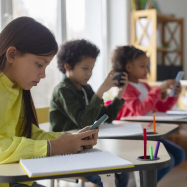 Diverse multiethnic kids students having break and using smartphones, sitting at desks in classroom, children playing games and checking social media at school