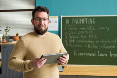Portrait of Caucasian man wearing glasses standing in classroom holding digital tablet smiling at camera with chalkboard behind displaying AI prompting instructions