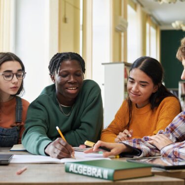 Group of students working on an algebra project in classroom setting, engaging in collaborative learning while sitting around table and sharing ideas on assignment