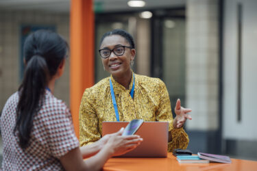 Two female teachers sitting at a table, having a discussion in the lobby area in the school they work at in Gateshead, North East England. They are talking about school issues together while using a laptop.